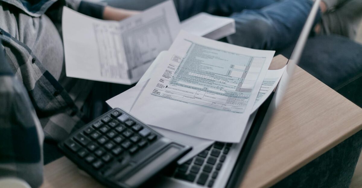 A young adult using a calculator and laptop to manage finances at home surrounded by papers