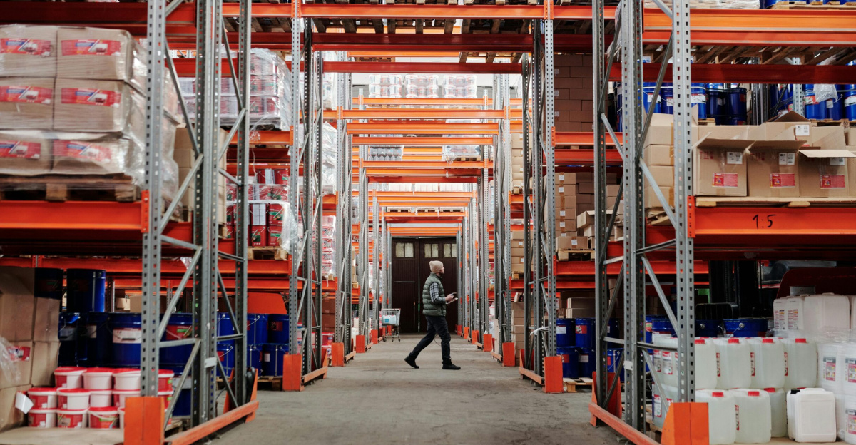 A man walking through a large industrial warehouse with stacked shelves filled with goods and products.
