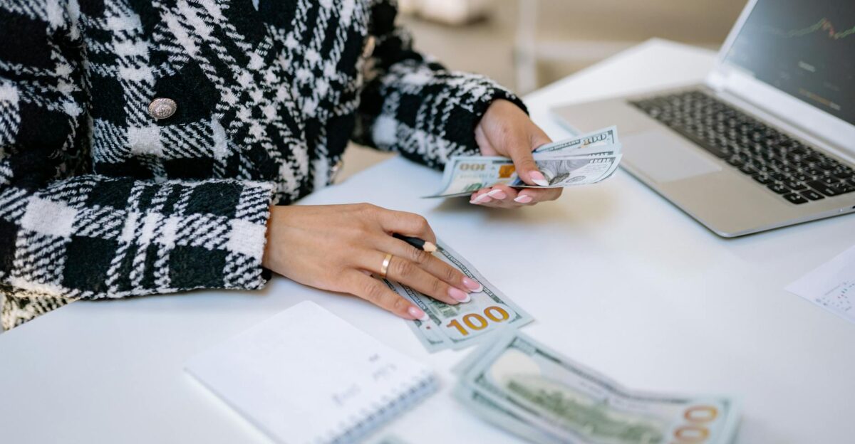 Close-up of a woman counting hundred-dollar bills at a desk with a laptop focusing on finance and work