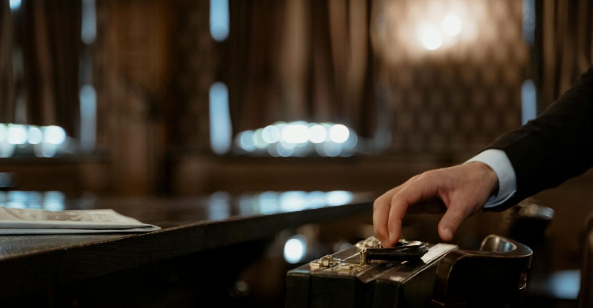 A close-up shot captures a hand holding a black briefcase in a dimly lit elegant room with a blurred background