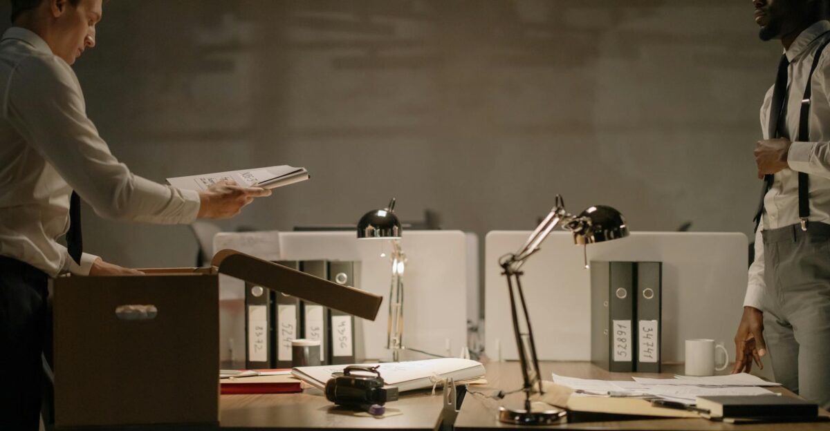 Two office workers sorting documents in a dimly lit workspace with several file binders